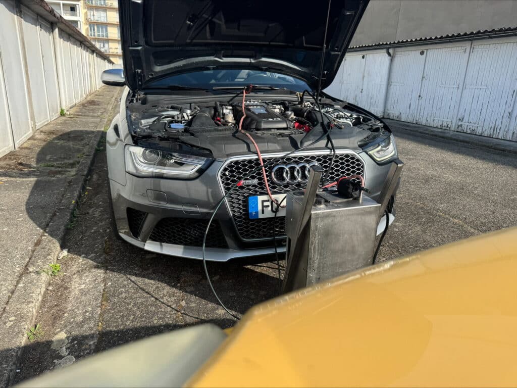 Audi RS4 gris capot ouvert, moteur exposé et câblé à un boîtier métallique pour réglage. Plaque française F, garages blancs en arrière-plan.